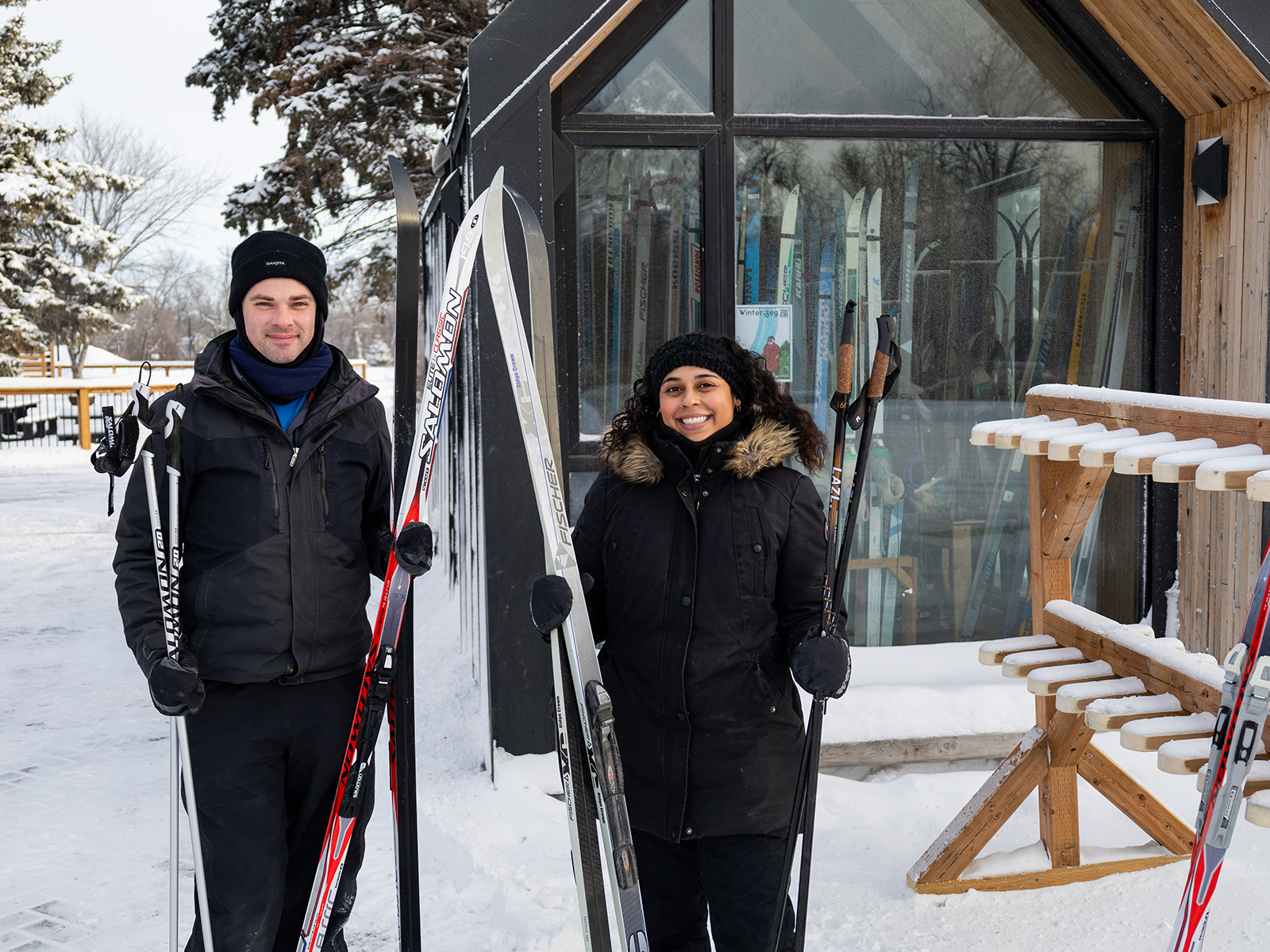 a man and a women smile at the camera holding cross country skis in front of a small rental shack